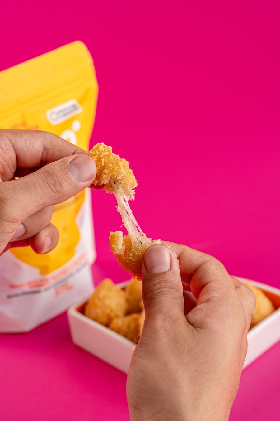 Mãos partindo uma mini coxinha Sucré com o queijo puxando um fio, sobre o fundo rosa da marca