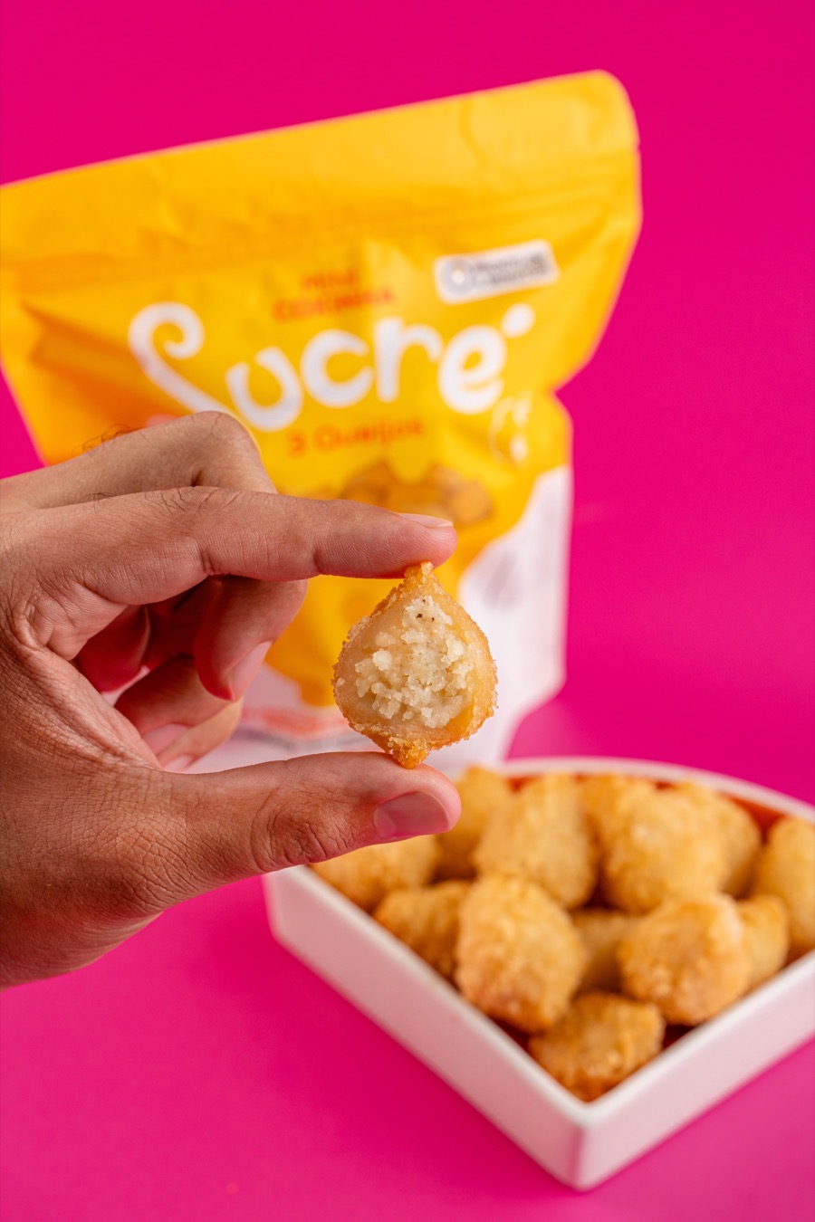 Mini coxinha 3 queijos mordida mostrando o recheio cremoso, com a embalagem amarela Sucré e uma tigela de coxinhas douradas ao fundo